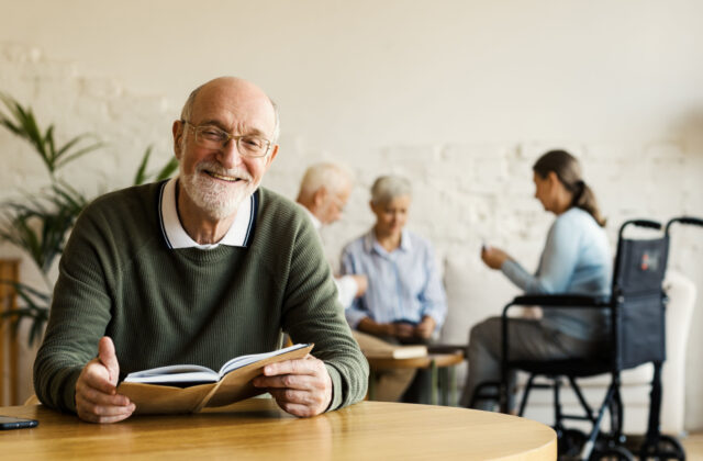 A senior man smiling while sitting at a table holding a book with other seniors in the background