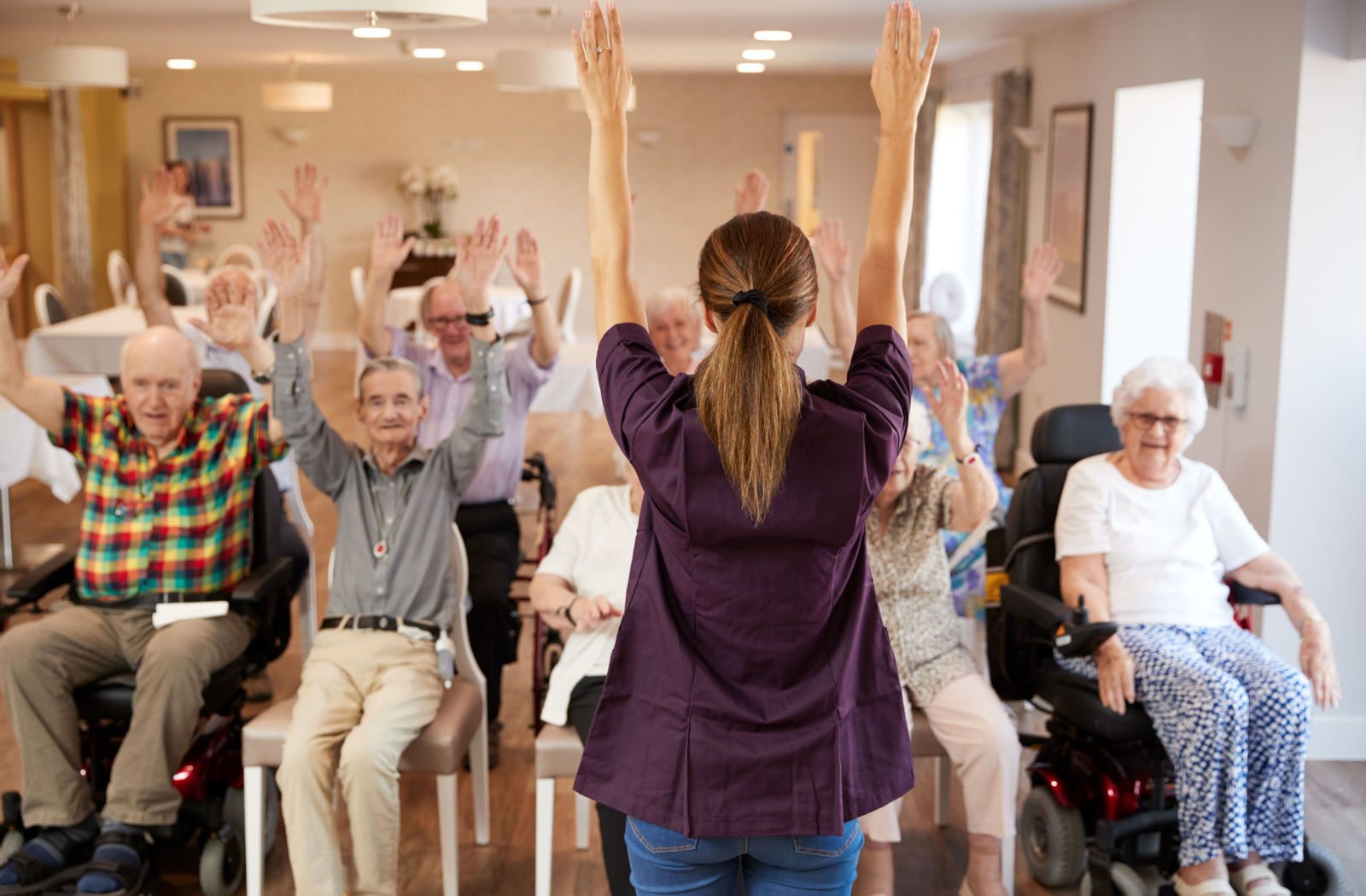 A group of seniors in an audience, each raising their right hand in a fist, some are smiling, as a nurse stands and leads the exercise