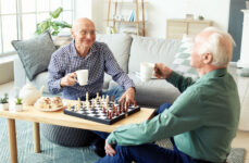 Two older adult men sitting at a coffee table and playing chess while sharing cinnamon rolls and a pot of tea
