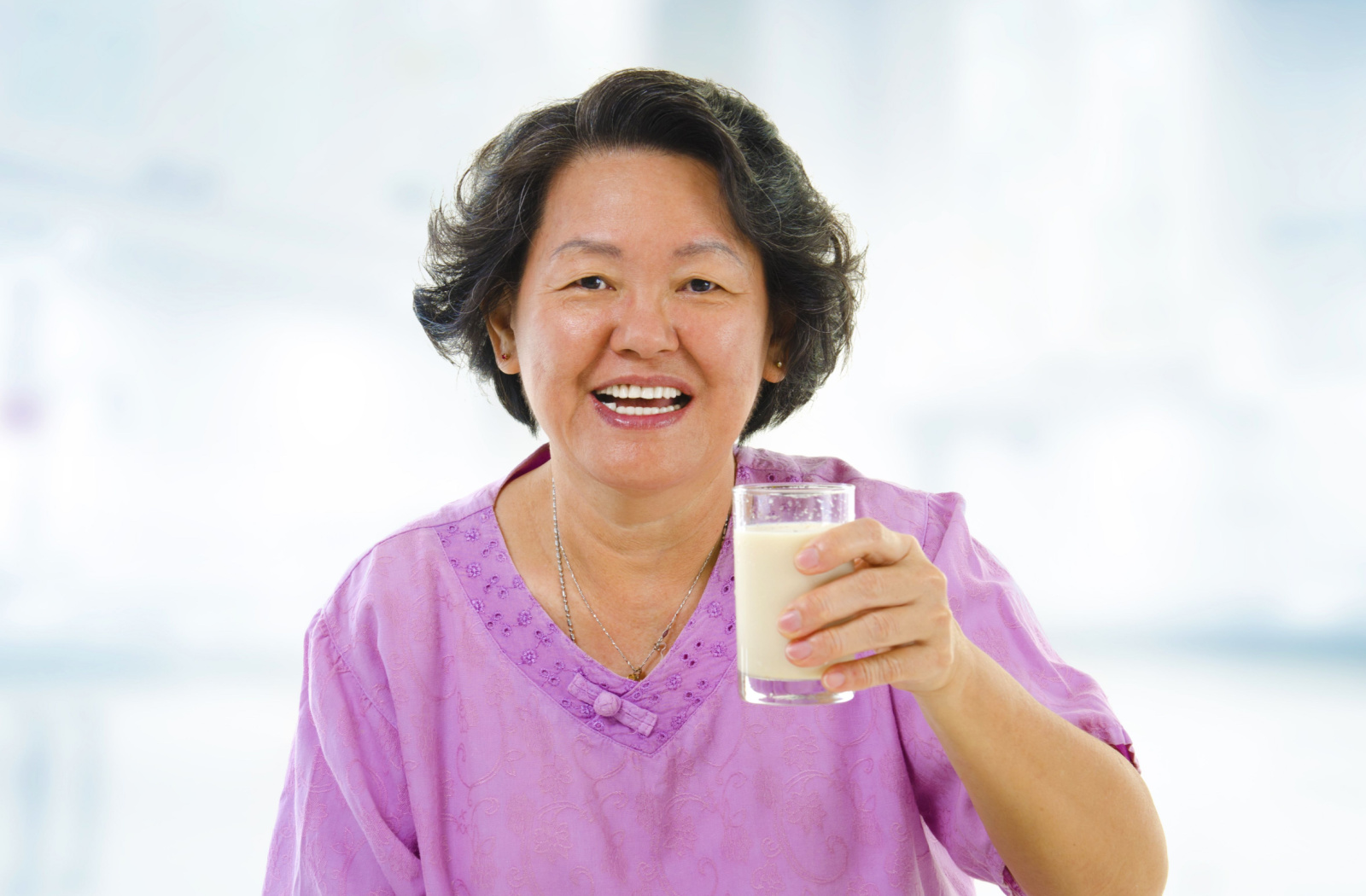 An older adult woman holding a glass of soy milk on her left hand, smiling and looking directly at the camera