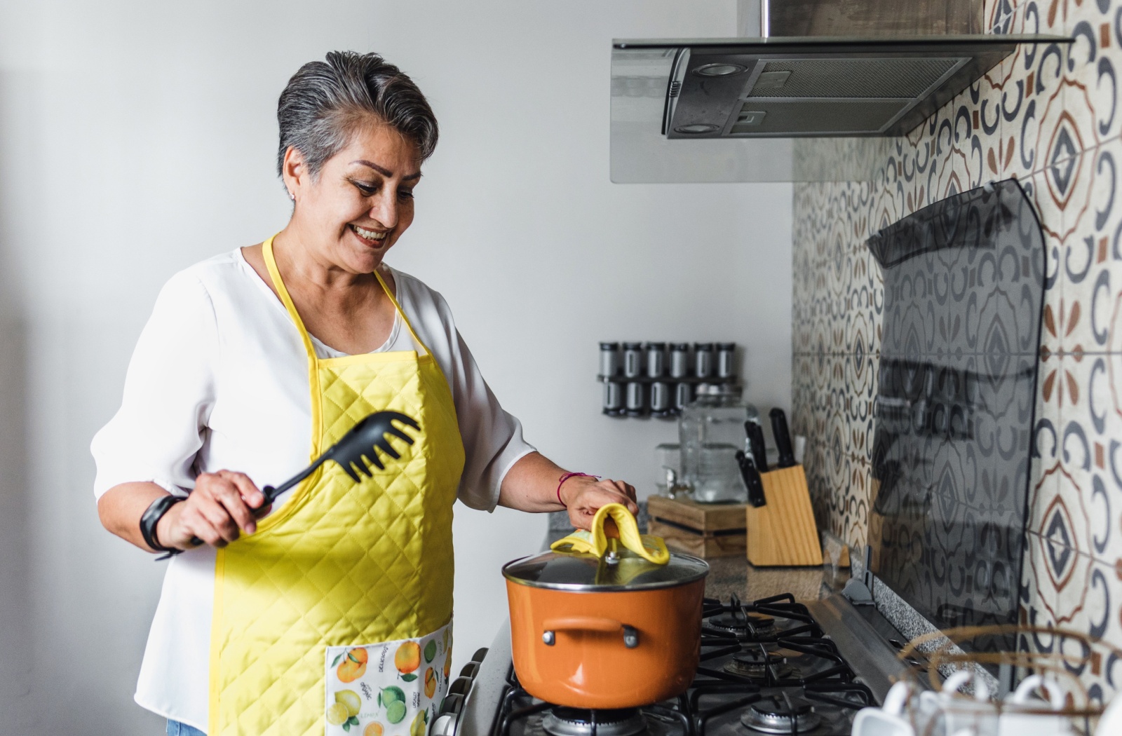A senior woman smiling and removing the lid of of a pot on a stove

